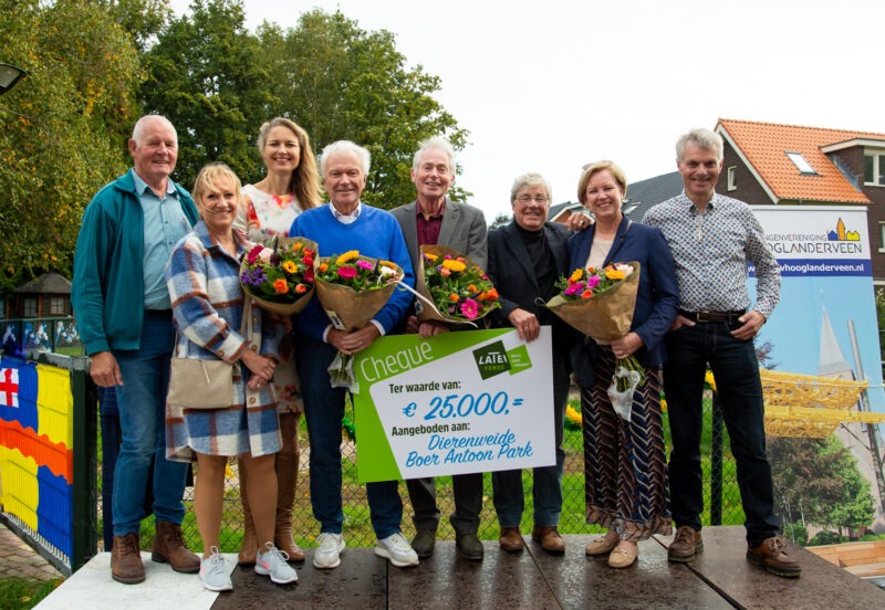 Een groep van acht lachende mensen staat buiten op een podium en houdt boeketten bloemen en een grote symbolische cheque van 25.000 euro vast, aangeboden aan Dierenweide Boer Antoon Park.