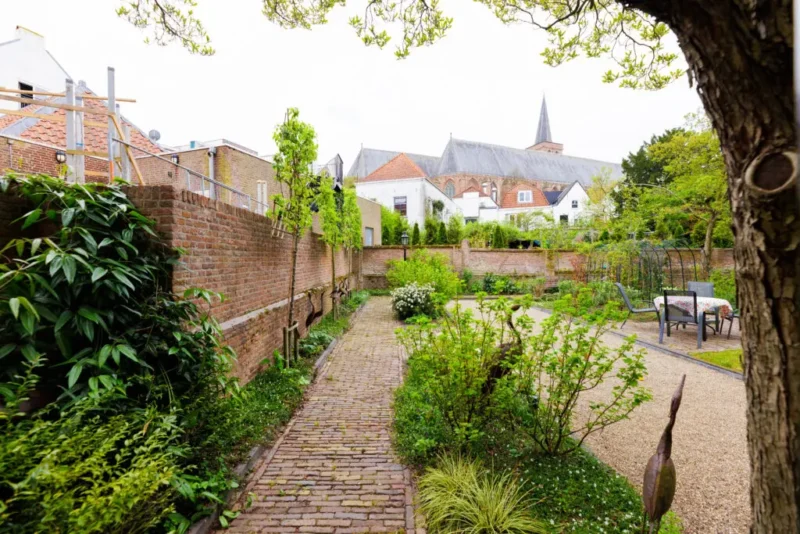 Een binnentuin met een oud klinkerpad en een jeu-de-boulesbaan, omringd door hoge bakstenen muren. Op de achtergrond zijn witte stadswoningen en de daken van een grote kerk zichtbaar.