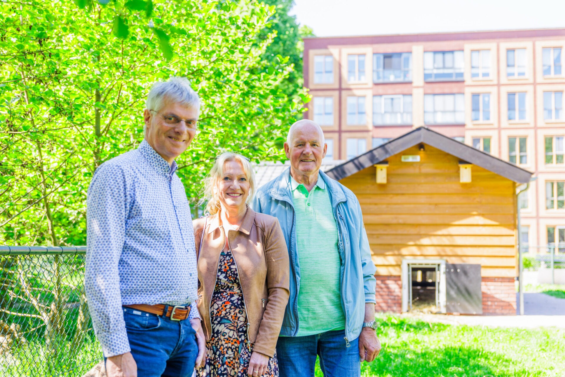 Een vrouw en twee mannen staan glimlachend buiten op een grasveld, met een houten dierenverblijf en een modern appartementencomplex op de achtergrond.