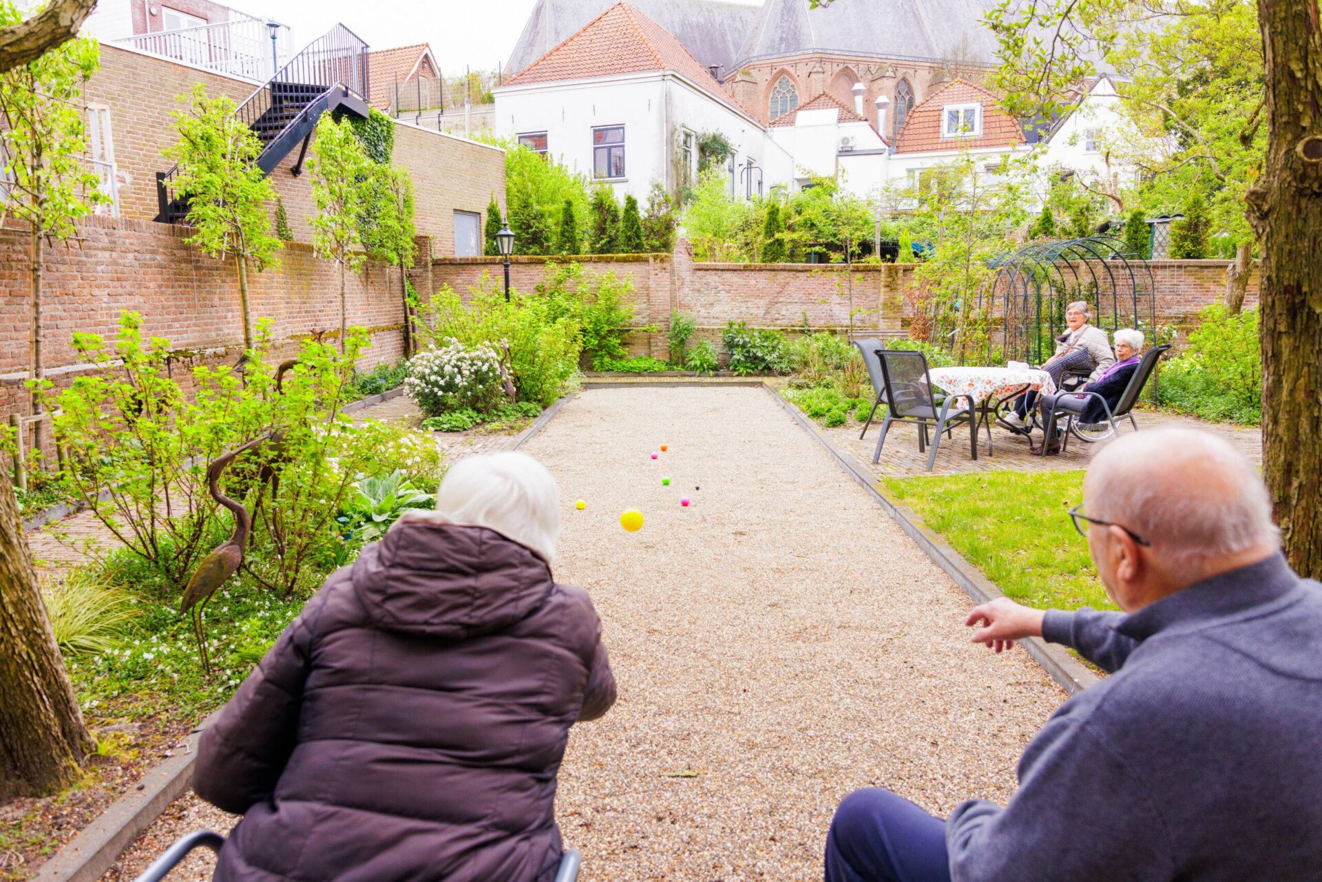 Gezien vanaf de rug spelen een oudere vrouw en man jeu de boules op een grindbaan in een binnentuin, terwijl twee andere senioren toekijken vanaf een terrasje.