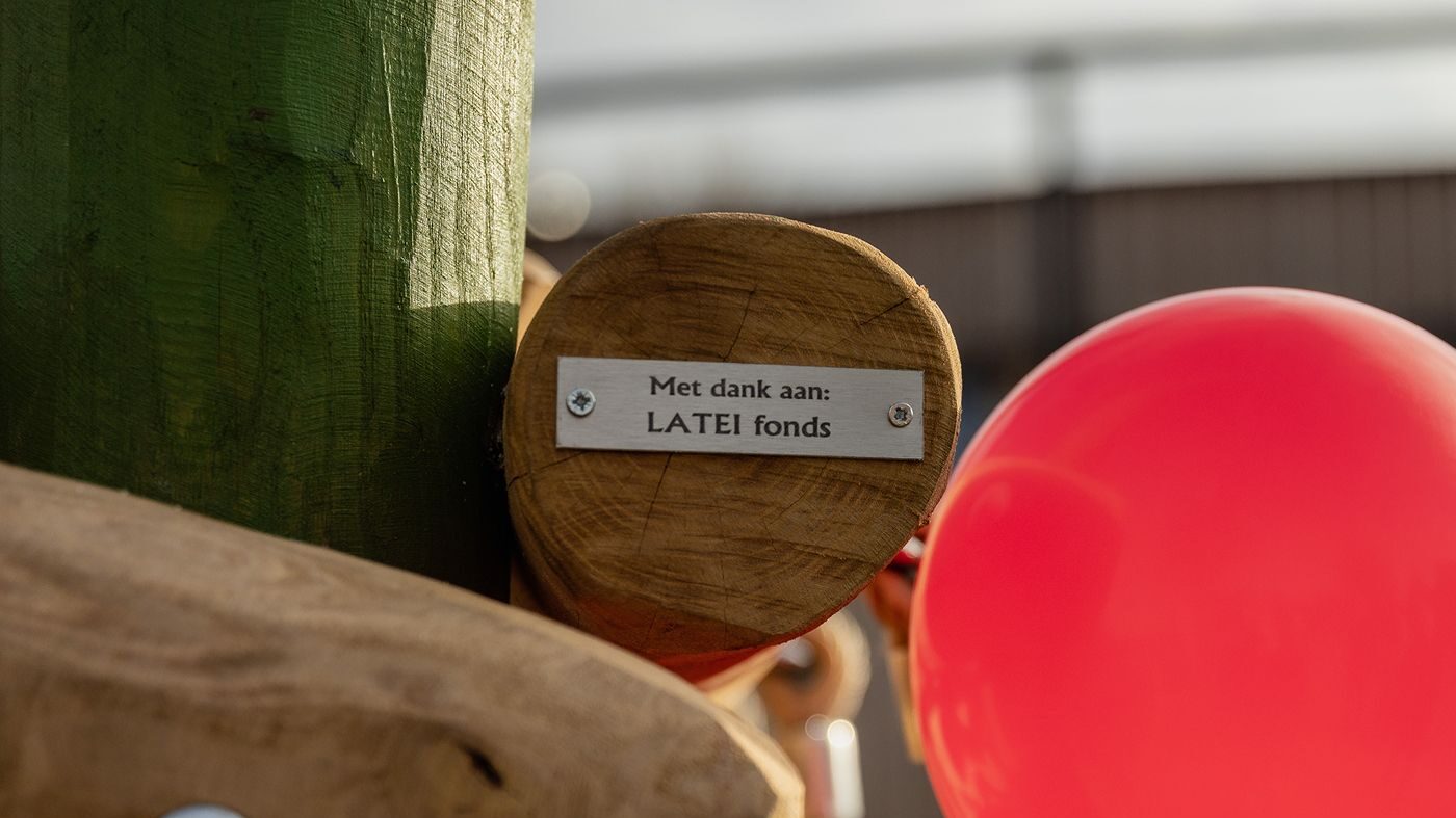 Close-up van een metalen plaatje op de kopse kant van een houten balk van een speeltoestel, met de tekst 'Met dank aan: LATEI fonds'. Rechts is een glimmende rode ballon zichtbaar.