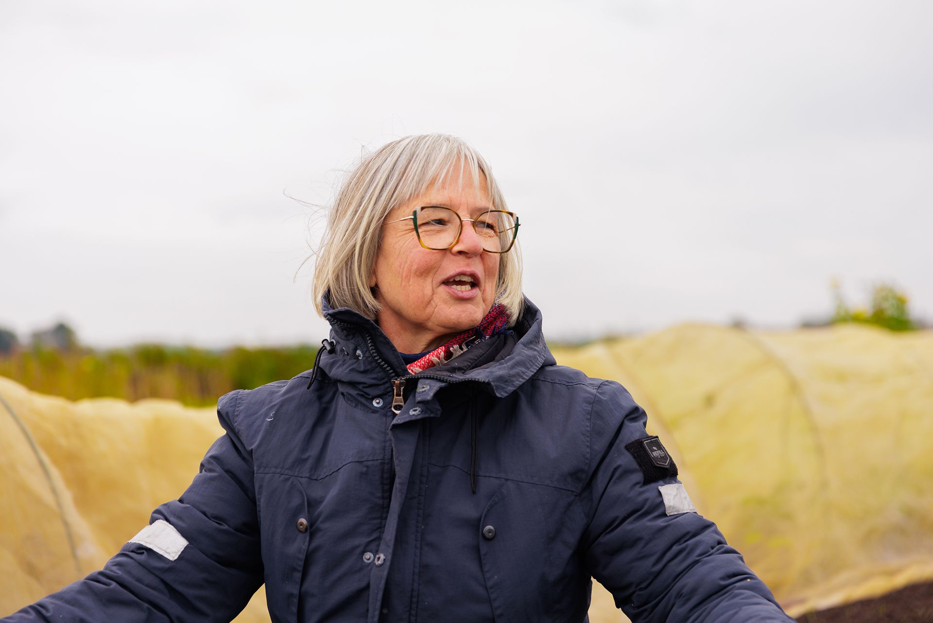 Een pratende vrouw met een bril en halflang grijs haar draagt een dikke, donkerblauwe winterjas in een buitenomgeving met gele landbouw- of kweeknetten op de achtergrond.