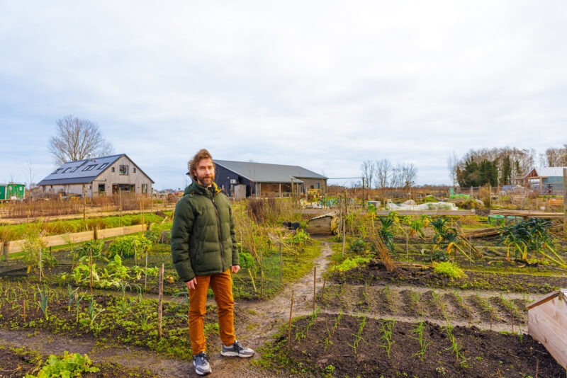 Een man met een baard in een winterjas staat op een zandpad in een grote moestuin. Op de achtergrond zijn houten schuren en landbouwvelden te zien onder een bewolkte lucht.