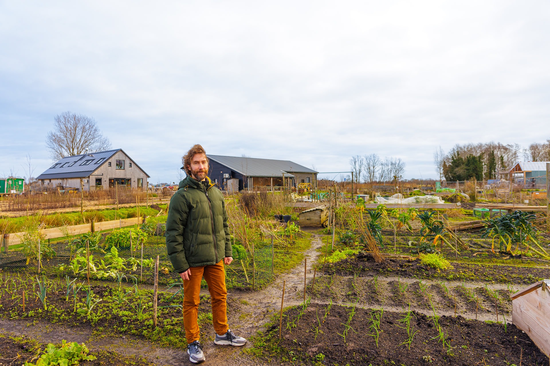 Een man met een baard in een winterjas staat op een zandpad in een grote moestuin. Op de achtergrond zijn houten schuren en landbouwvelden te zien onder een bewolkte lucht.