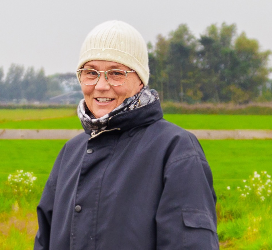 Een glimlachende vrouw met een bril, een witte muts en een donkerblauwe winterjas staat buiten voor een weids groen weiland.