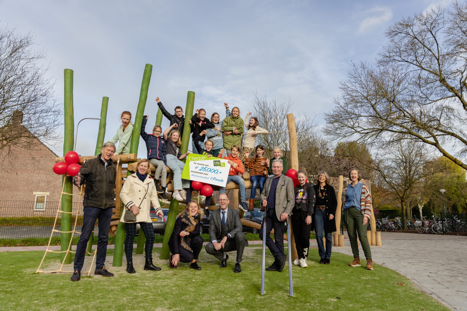 Een groep enthousiaste kinderen zit op een groot houten speeltoestel en houdt een symbolische cheque van 25.000 euro vast, omringd door glimlachende volwassenen in een buitenspeeltuin.