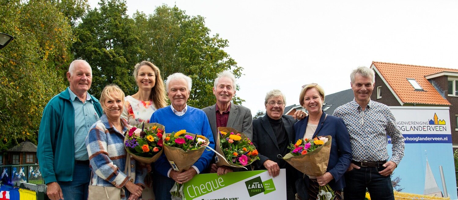 Een groep glimlachende mensen staat buiten naast elkaar en houdt kleurrijke boeketten bloemen en een symbolische cheque van het Latei Fonds vast. Rechts staat een banner van Belangenvereniging Hooglanderveen.