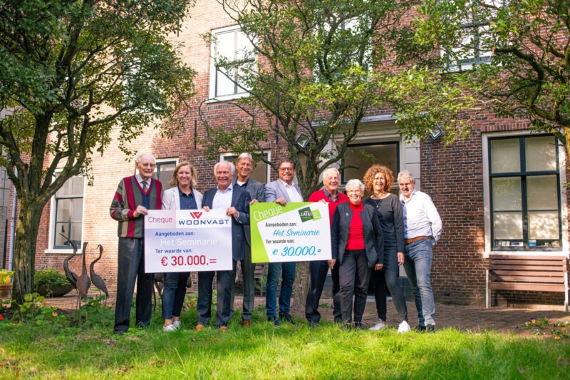 Een groep mensen staat in een tuin voor een historisch bakstenen gebouw en toont twee grote symbolische cheques van elk 30.000 euro (van Woonvast en het Latei Fonds), beide aangeboden aan 'Het Seminarie'.