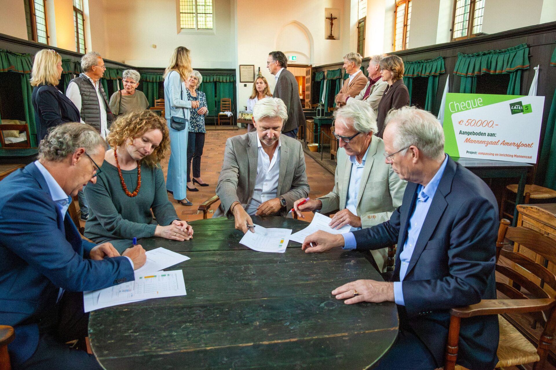 Een groep mensen in een historische zaal, waarbij vijf personen aan een grote ronde houten tafel documenten bestuderen en ondertekenen. Op de achtergrond staat een grote symbolische cheque van 50.000 euro voor de Mannenzaal Amersfoort.