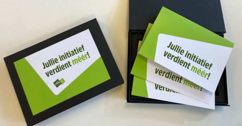 Een zwarte geschenkdoos met promotiemateriaal van het Latei Fonds. Op de gesloten doos en op de kaarten in de geopende doos staat de tekst 'Jullie initiatief verdient méér!' op een groen-witte achtergrond.