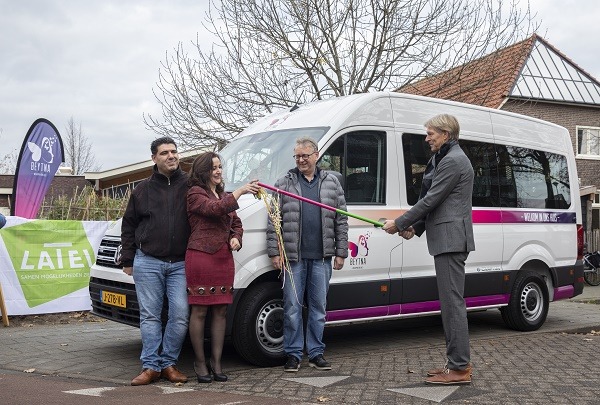Vier mensen staan buiten bij een witte personenbus van 'Beytna'. Een man in een grijs pak overhandigt een grote, feestelijk versierde sleutel aan de groep. Op de achtergrond is een spandoek van het Latei Fonds zichtbaar.