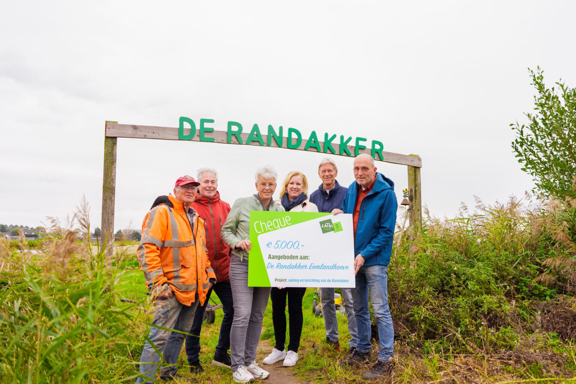 Een groep van zes mensen poseert buiten onder een houten poort met de tekst 'De Randakker' en houdt een symbolische cheque van 5.000 euro vast voor De Randakker Eemlandhoeve.