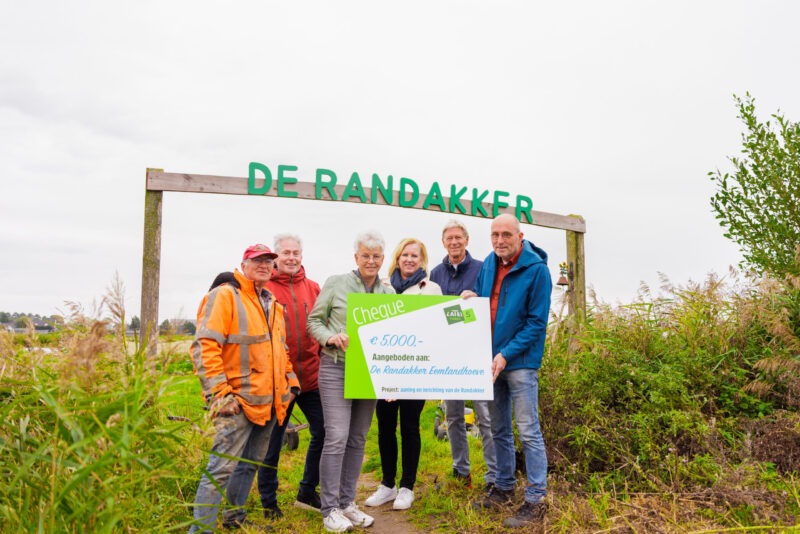 Een groep van zes mensen poseert buiten onder een houten poort met de tekst 'De Randakker' en houdt een symbolische cheque van 5.000 euro vast voor De Randakker Eemlandhoeve.