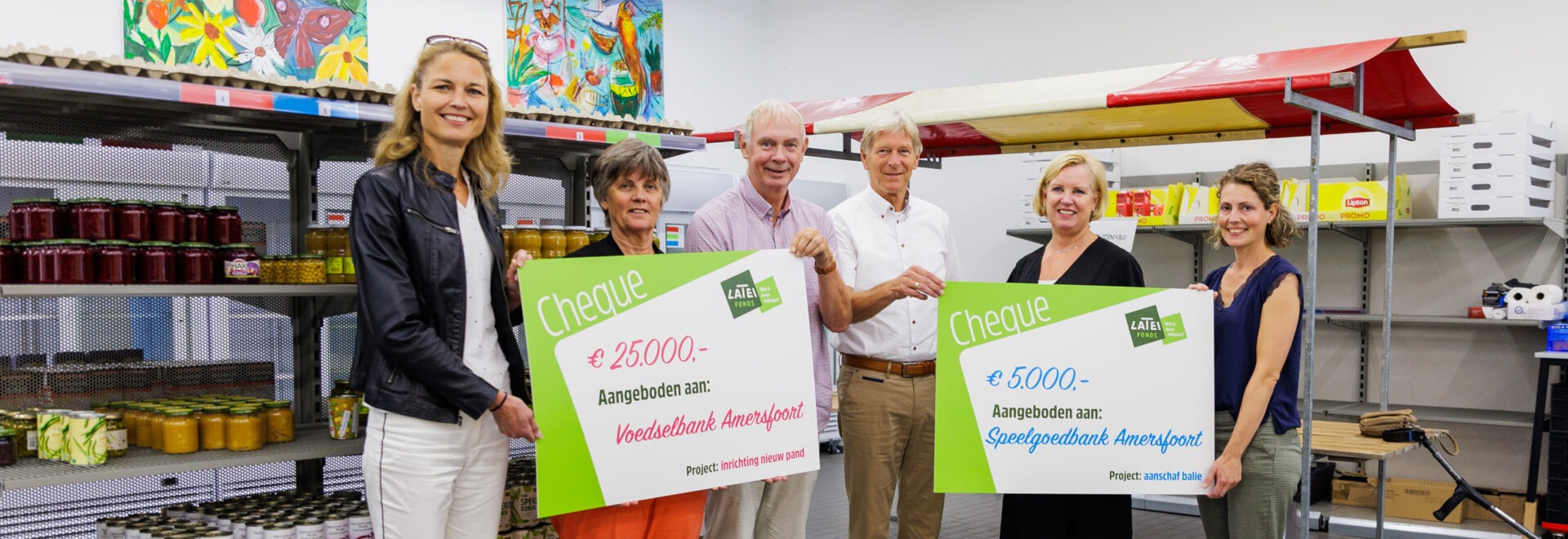 Een groep van zes personen staat in een magazijnruimte en toont twee grote symbolische cheques van het Latei Fonds: één van 25.000 euro voor Voedselbank Amersfoort en één van 5.000 euro voor Speelgoedbank Amersfoort.
