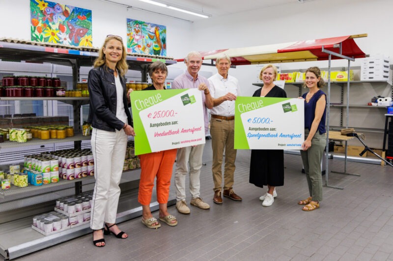 Een groep van zes personen staat in een magazijnruimte en toont twee grote symbolische cheques van het Latei Fonds: één van 25.000 euro voor Voedselbank Amersfoort en één van 5.000 euro voor Speelgoedbank Amersfoort.
