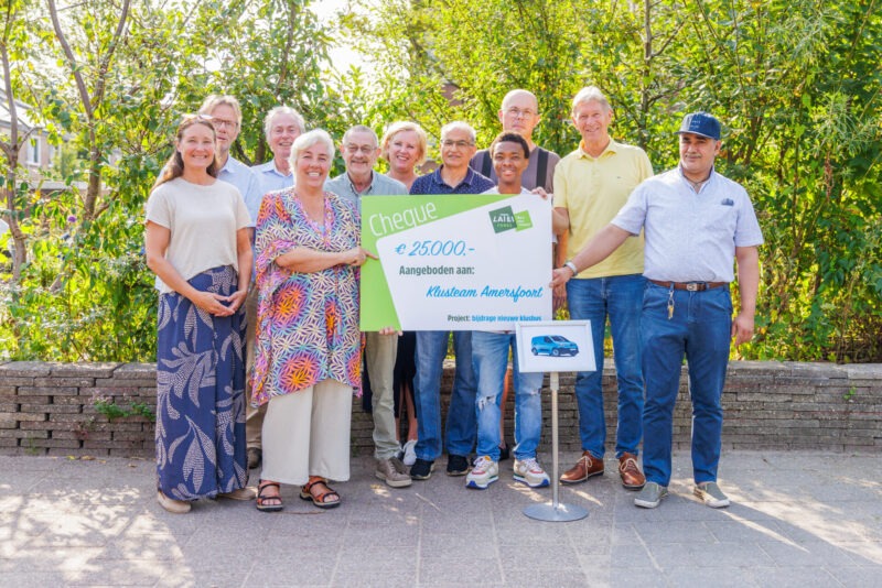 Een groep glimlachende mensen staat buiten voor een groene achtergrond en houdt een grote symbolische cheque van 25.000 euro vast, aangeboden aan Klusteam Amersfoort voor een nieuwe klusbus.