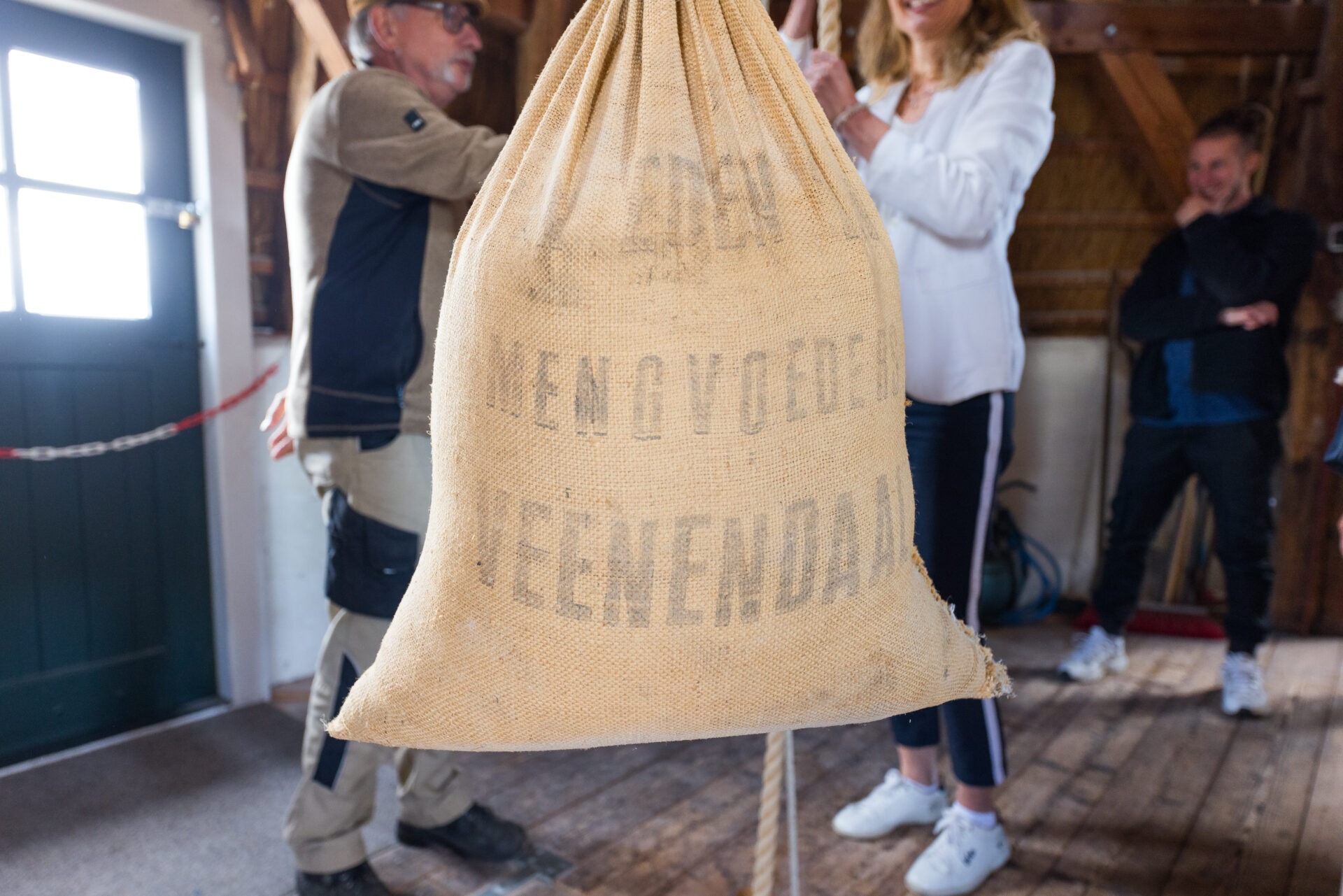Een grote, gevulde jute zak hangt centraal aan een touw in het houten interieur van een molen. Op de zak staat de vervaagde tekst 'MENGVOEDERS VEENENDAAL'. Op de achtergrond staan enkele personen toe te kijken.