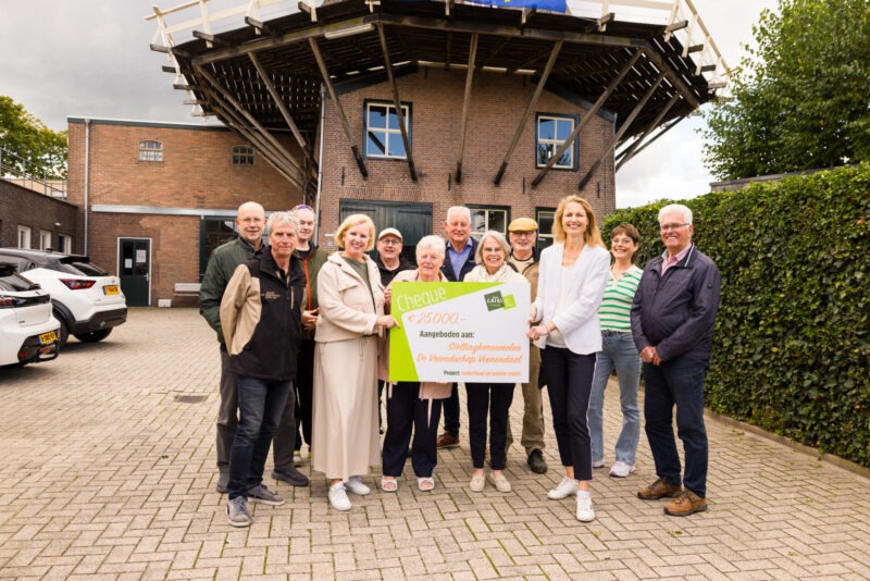 Een groep mensen staat buiten voor Stellingkorenmolen 'De Vriendschap' in Veenendaal en toont een grote symbolische cheque van het Latei Fonds ter waarde van 25.000 euro.
