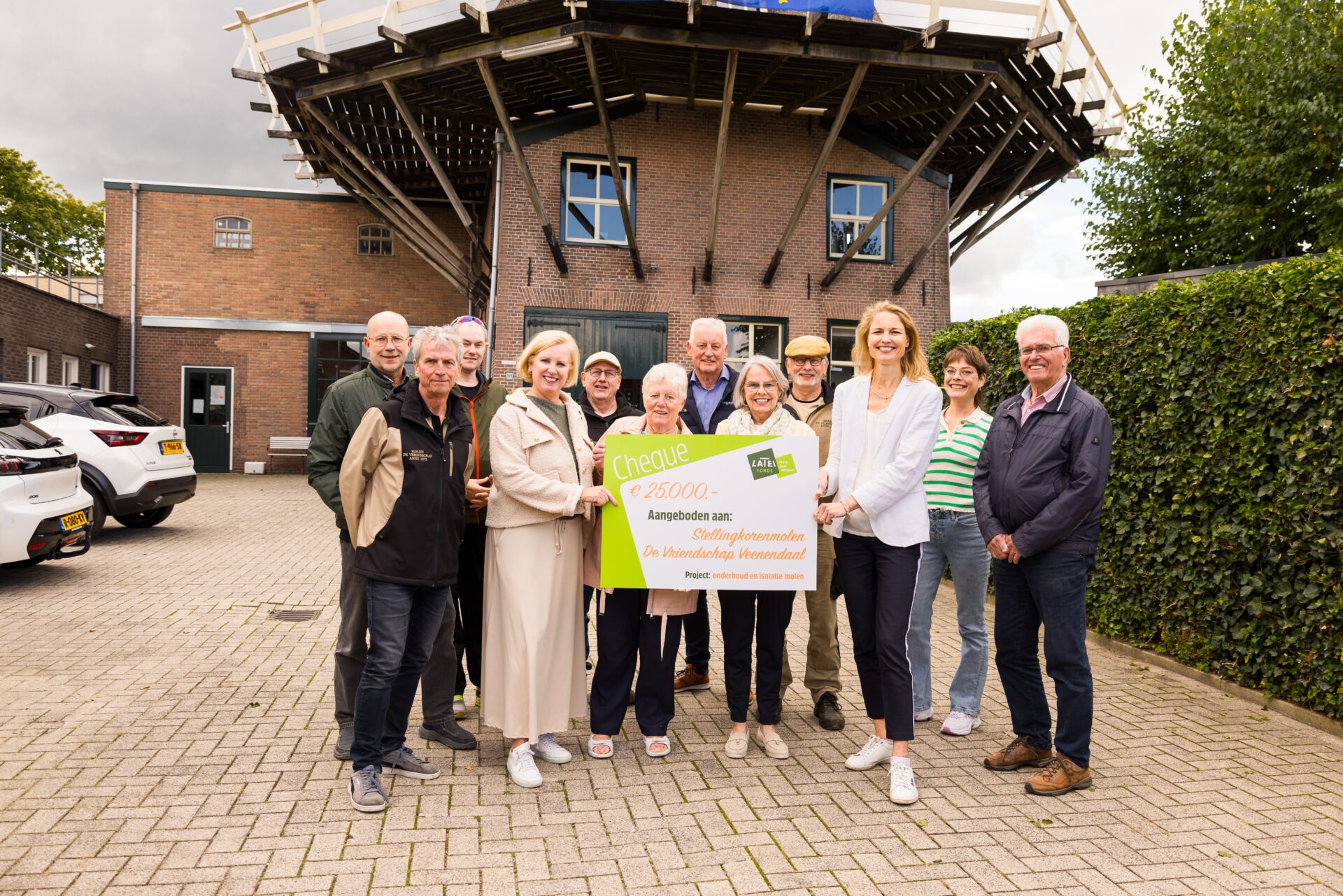 Een groep mensen staat buiten voor Stellingkorenmolen 'De Vriendschap' in Veenendaal en toont een grote symbolische cheque van het Latei Fonds ter waarde van 25.000 euro.