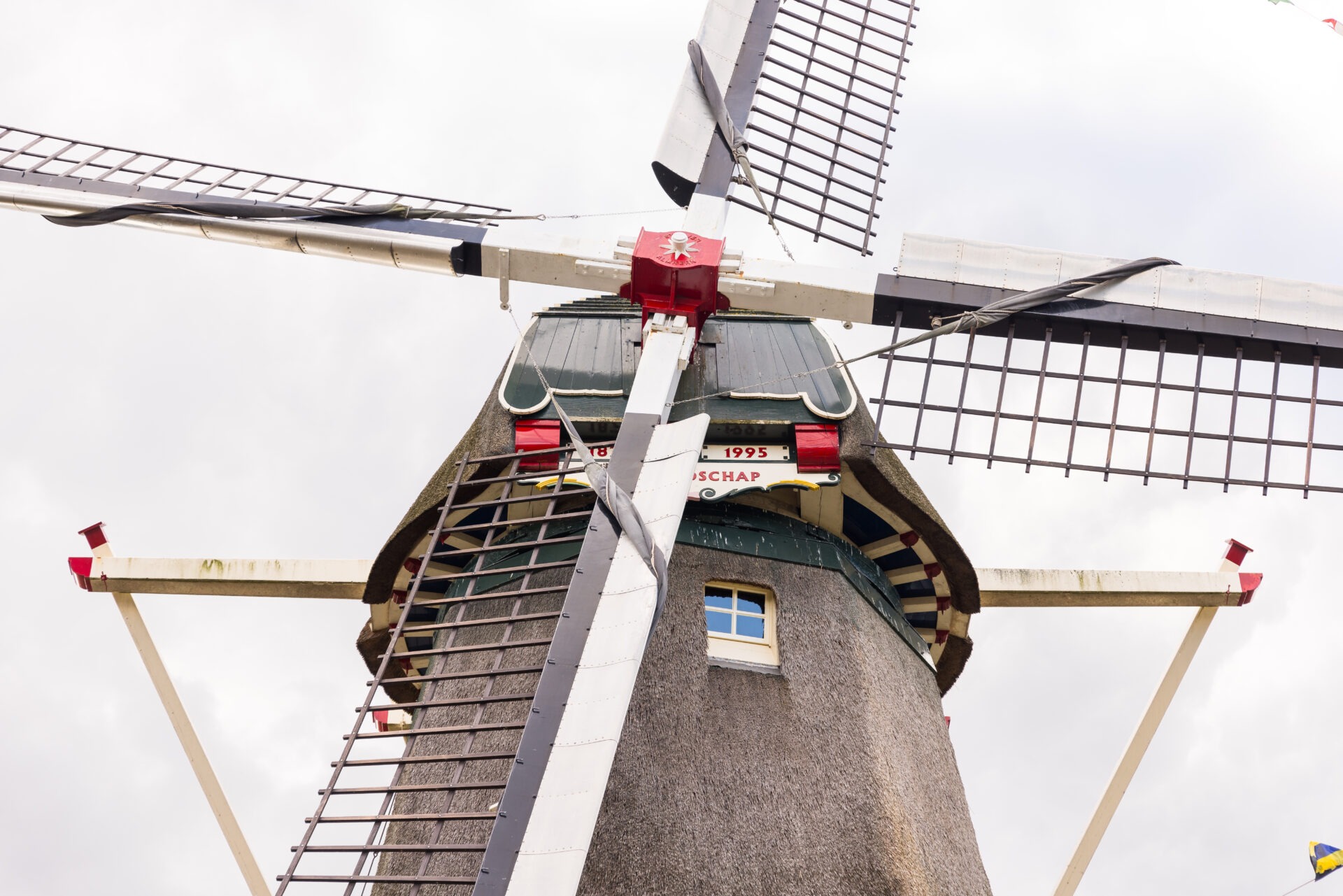 Een kikvorsperspectief van de bovenkant van korenmolen 'De Vriendschap', met de focus op de askop, de wieken en de rietgedekte kap tegen een bewolkte lucht.