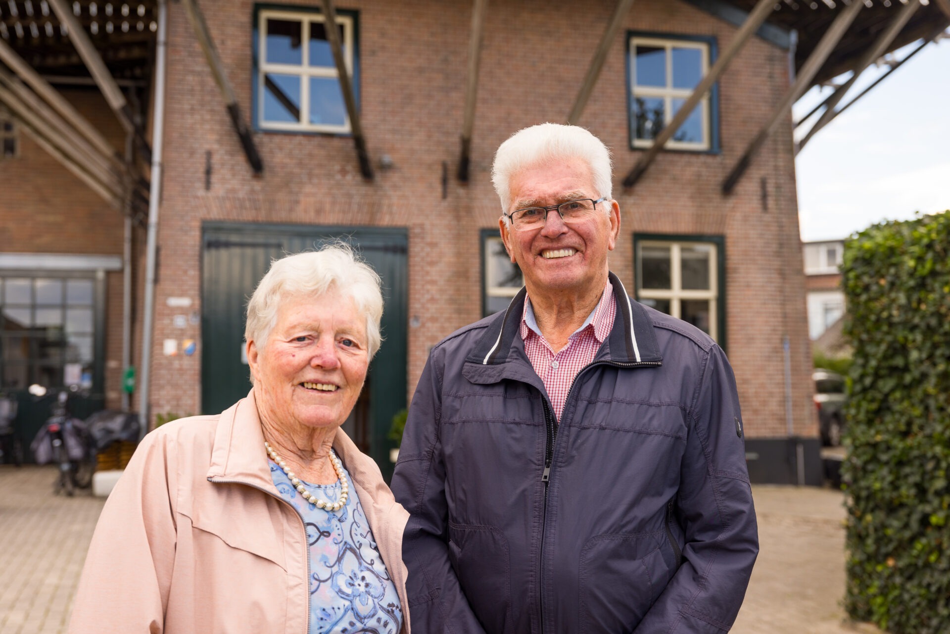 Een glimlachend ouder echtpaar staat samen buiten op de voorgrond, met de bakstenen onderbouw van een traditionele molen als achtergrond.