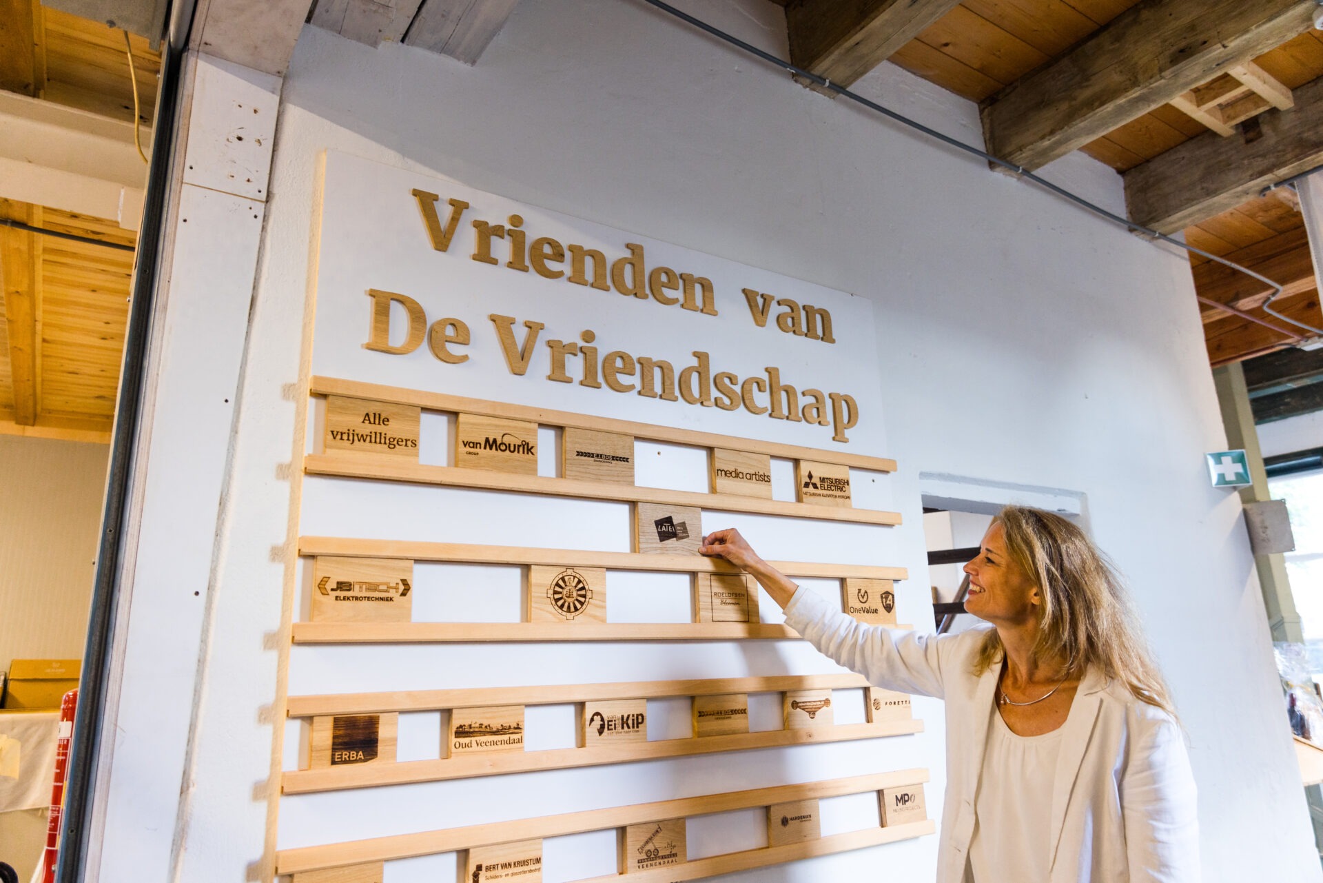 Een glimlachende vrouw plaatst een houten naambordje op een sponsorwand met de titel 'Vrienden van De Vriendschap' in een ruimte met een houten balkenplafond.