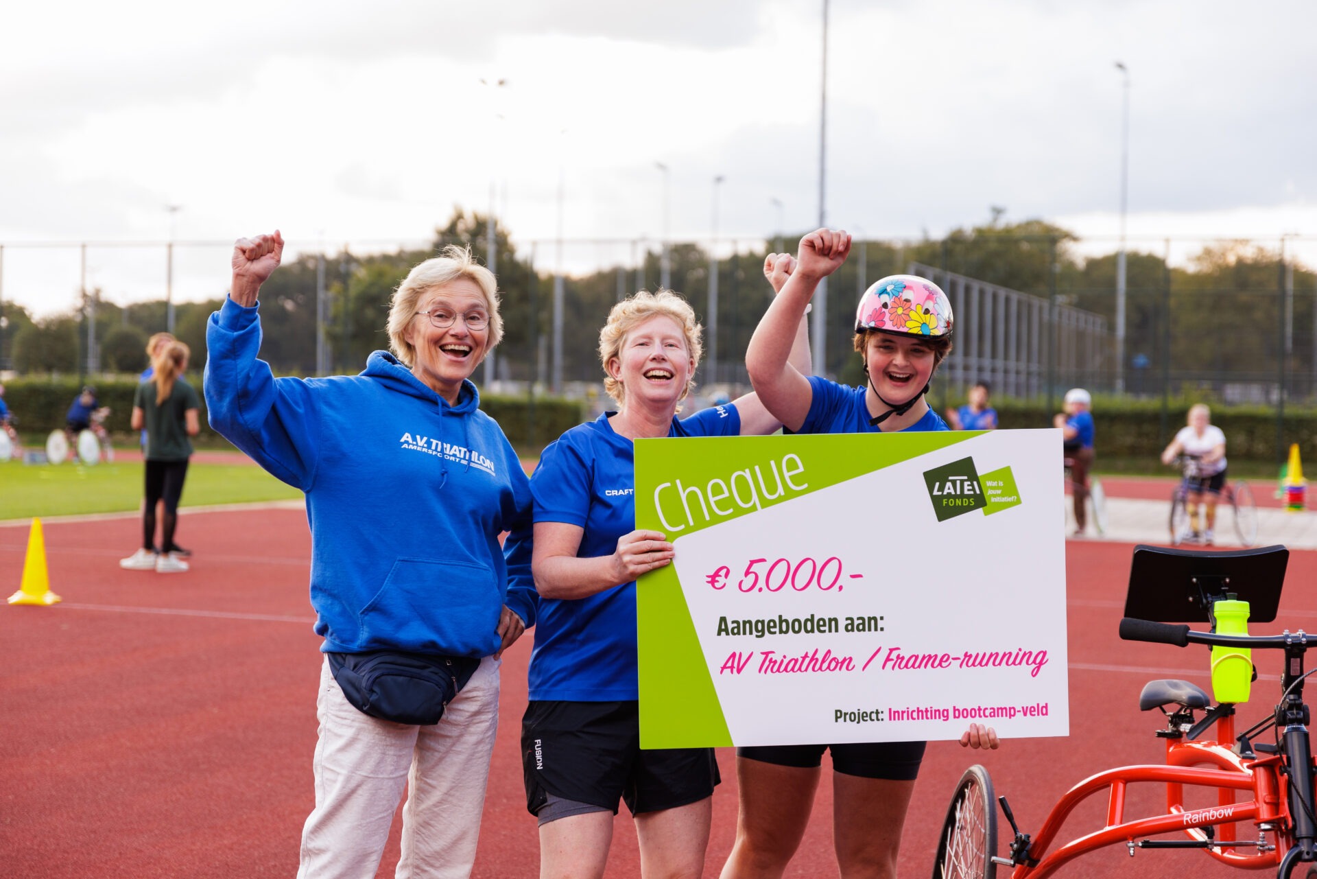 Drie enthousiaste vrouwen vieren feest op een atletiekbaan terwijl ze een grote symbolische cheque van 5.000 euro vasthouden voor 'AV Triathlon / Frame-running'.