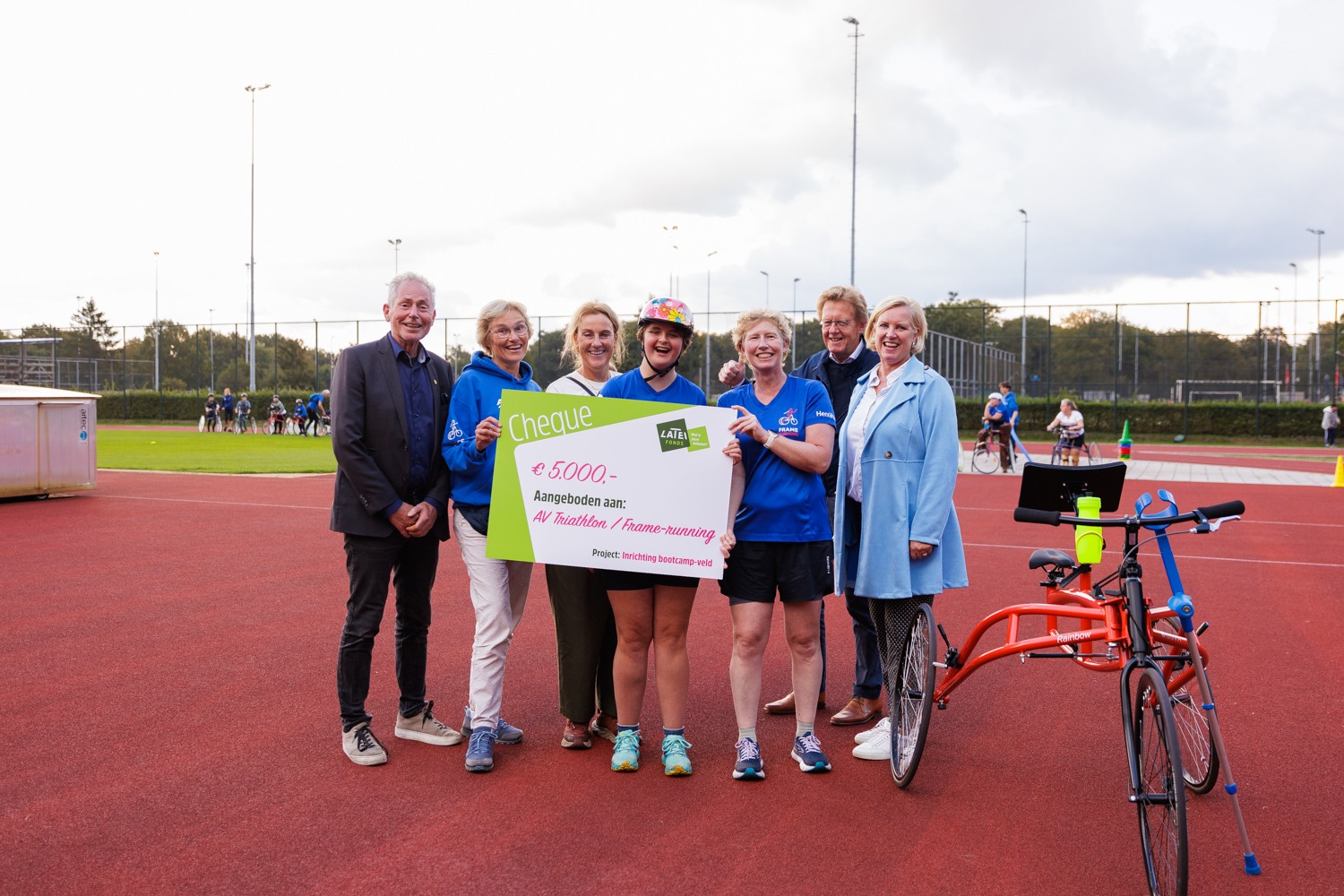 Een groep glimlachende mensen staat op een rode atletiekbaan en houdt een grote symbolische cheque vast van 5.000 euro voor 'AV Triathlon / Frame-running'. Rechts op de voorgrond staat een rode running frame (race runner) driewieler.