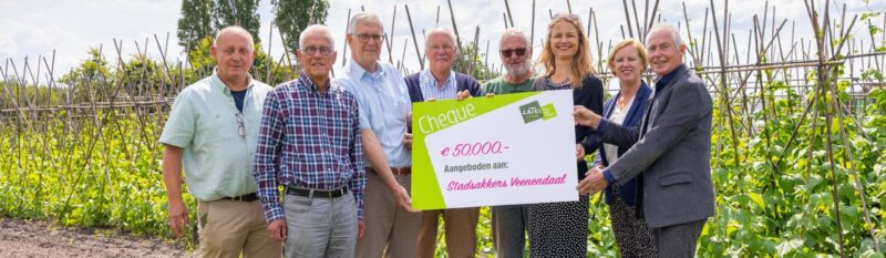 Een groep van acht mensen staat op een akker tussen de gewassen en houdt een grote symbolische cheque van 50.000 euro vast, aangeboden aan Stadsakkers Veenendaal.