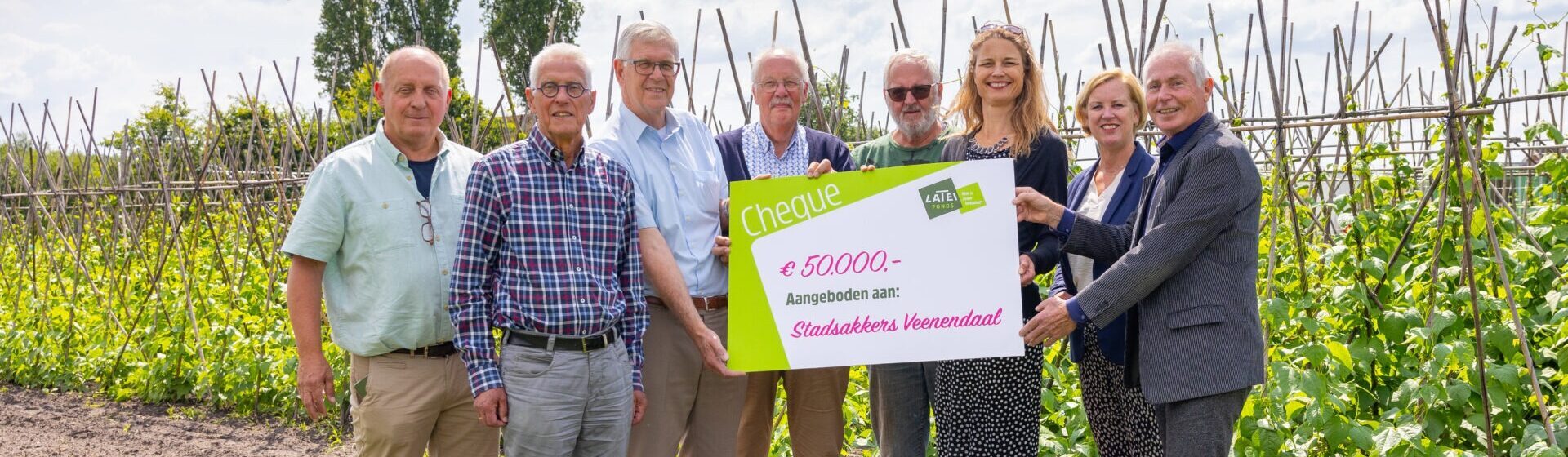 Een groep van acht mensen staat op een akker tussen de gewassen en houdt een grote symbolische cheque van 50.000 euro vast, aangeboden aan Stadsakkers Veenendaal.