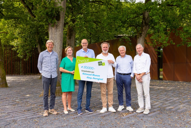 Een groep van zes personen staat buiten tussen grote bomen en houdt een grote symbolische cheque van 50.000 euro vast, aangeboden aan Nationaal Museum Kamp Amersfoort.