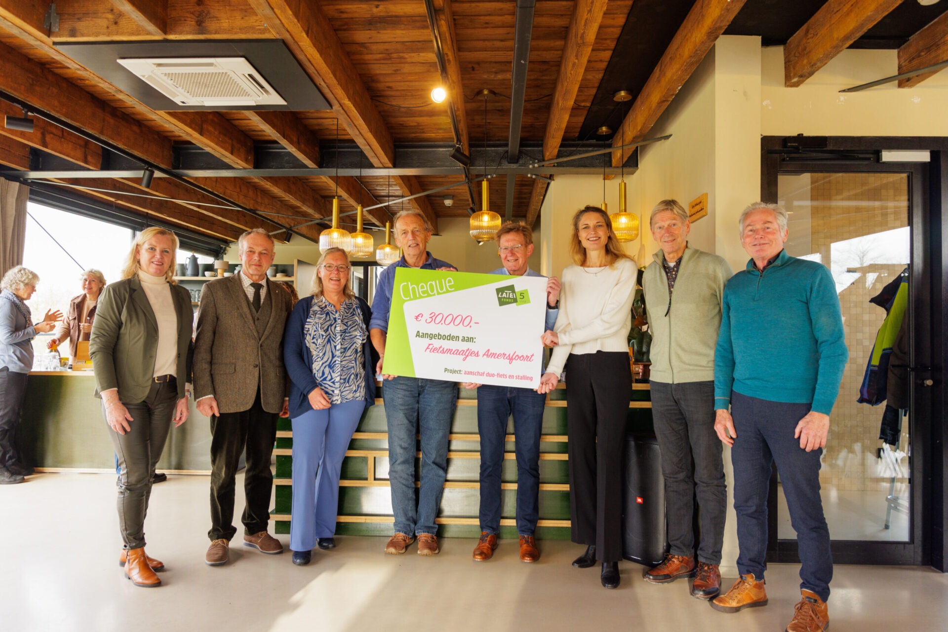 Een groep van zeven mensen poseert binnen in een modern gebouw met een grote symbolische cheque van 30.000 euro van het Latei Fonds voor Fietsmaatjes Amersfoort.