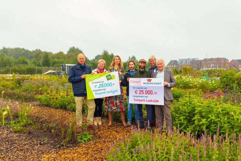 Een groep mensen staat in een kleurrijke bloementuin en toont twee grote symbolische cheques van elk 25.000 euro (van het Latei Fonds en Woonvast), beide aangeboden aan Tuinpark Laakzijde.