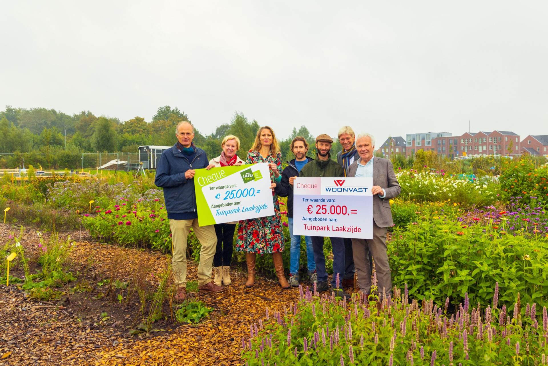 Een groep mensen staat in een kleurrijke bloementuin en toont twee grote symbolische cheques van elk 25.000 euro (van het Latei Fonds en Woonvast), beide aangeboden aan Tuinpark Laakzijde.