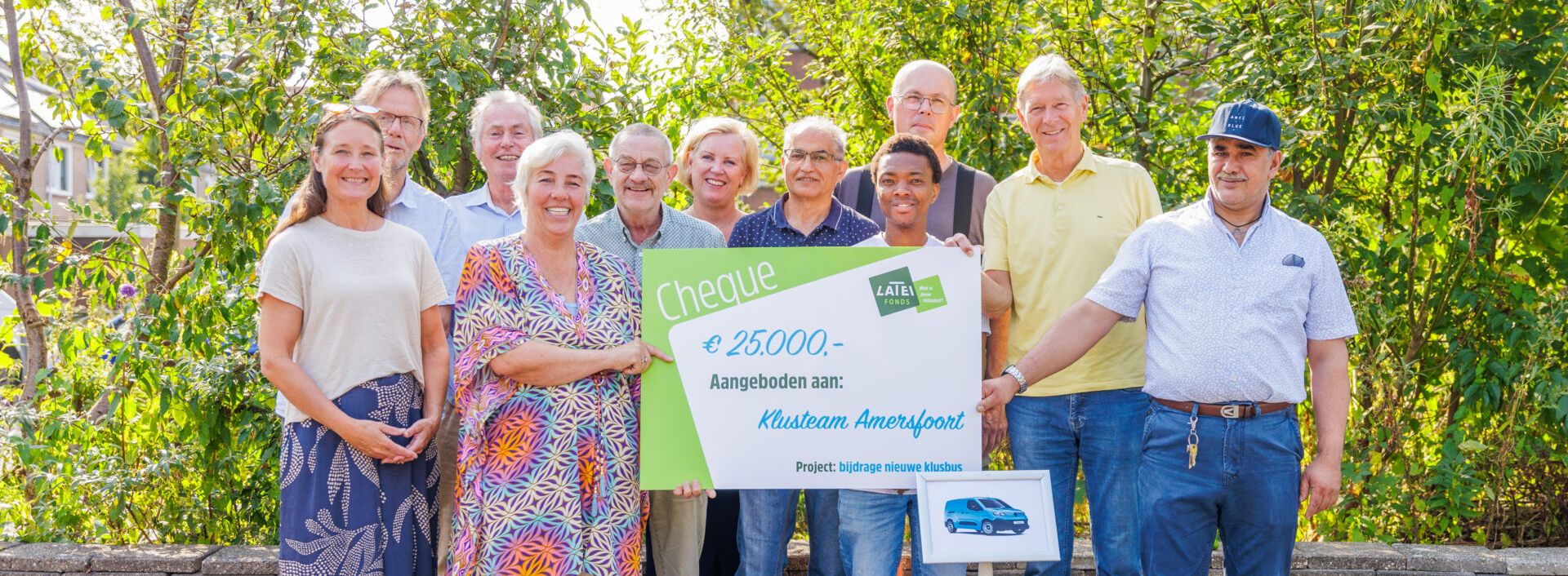 Een groep glimlachende mensen staat buiten voor een groene achtergrond en houdt een grote symbolische cheque van 25.000 euro vast, aangeboden aan Klusteam Amersfoort voor een nieuwe klusbus.