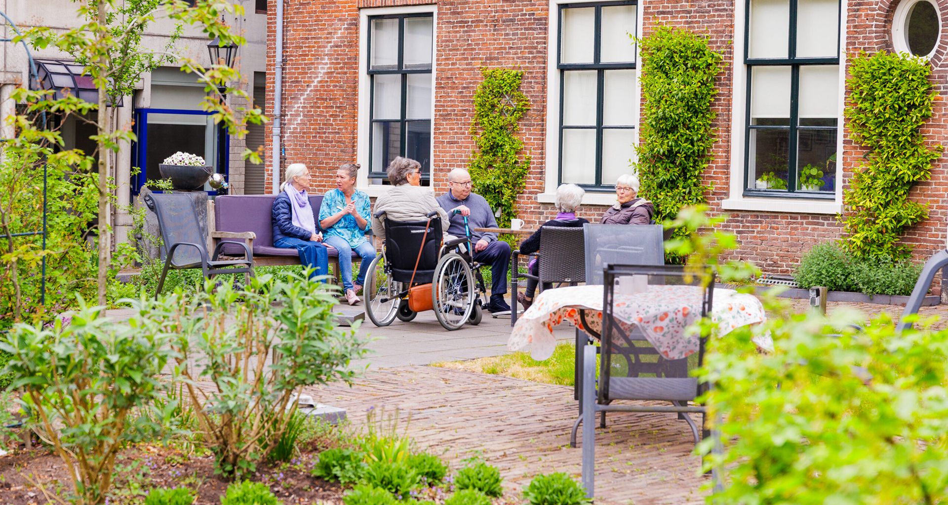 Een groep ouderen en een zorgmedewerker zitten gezellig samen in een groene, beschutte binnentuin bij een historisch bakstenen gebouw.