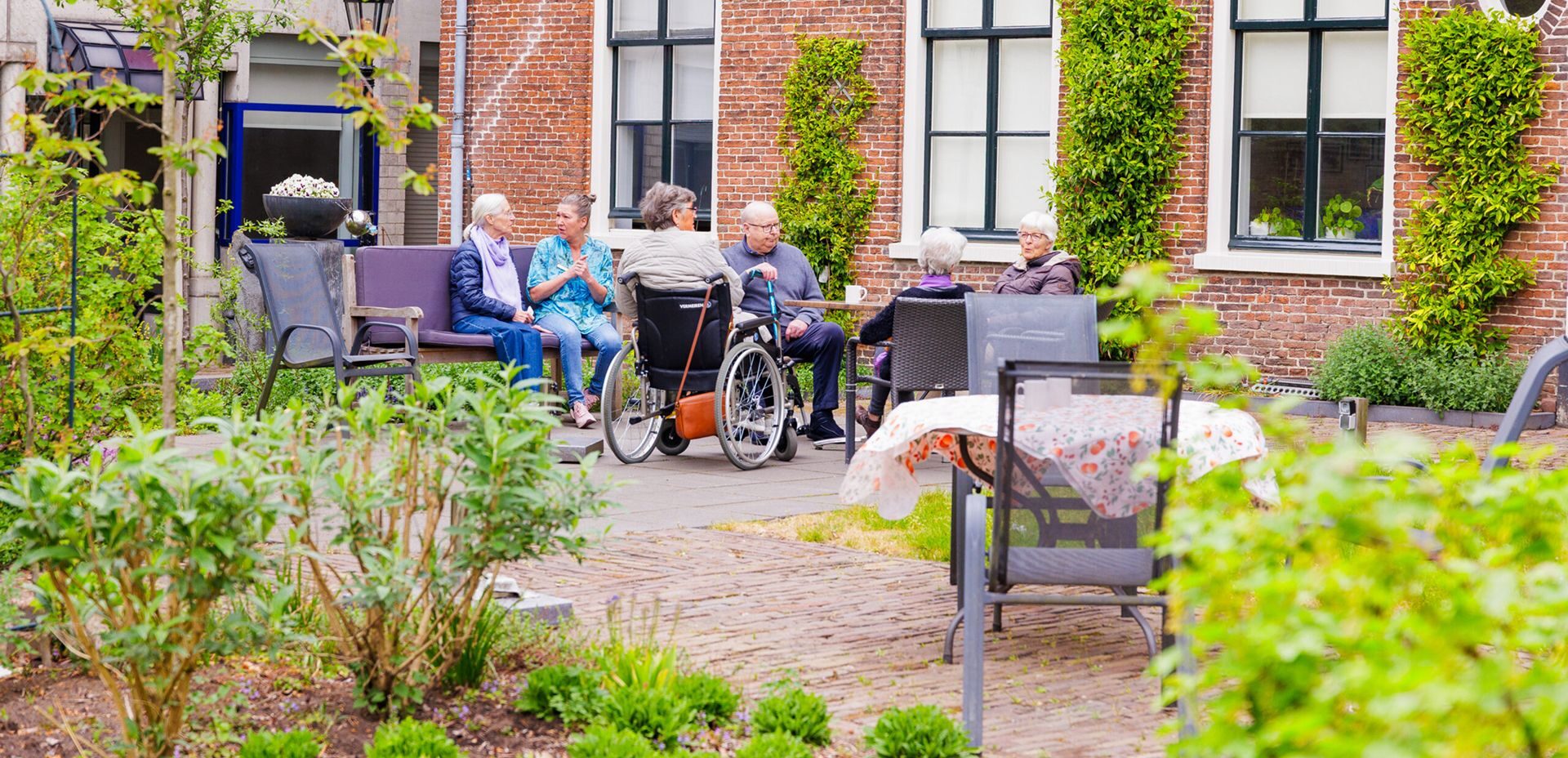 Een groep ouderen en een zorgmedewerker zitten gezellig samen in een groene, beschutte binnentuin bij een historisch bakstenen gebouw.