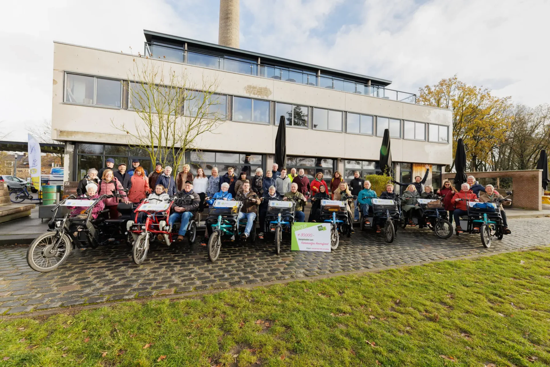 Een grote groep mensen poseert buiten voor een modern gebouw met meerdere duofietsen en een symbolische cheque van 30.000 euro voor Fietsmaatjes Amersfoort.