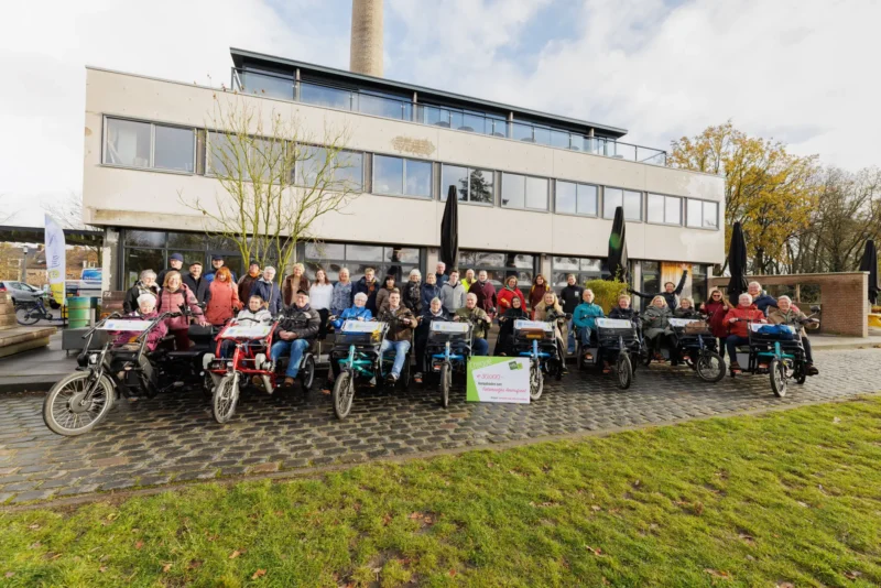 Een grote groep mensen poseert buiten voor een modern gebouw met meerdere duofietsen en een symbolische cheque van 30.000 euro voor Fietsmaatjes Amersfoort.