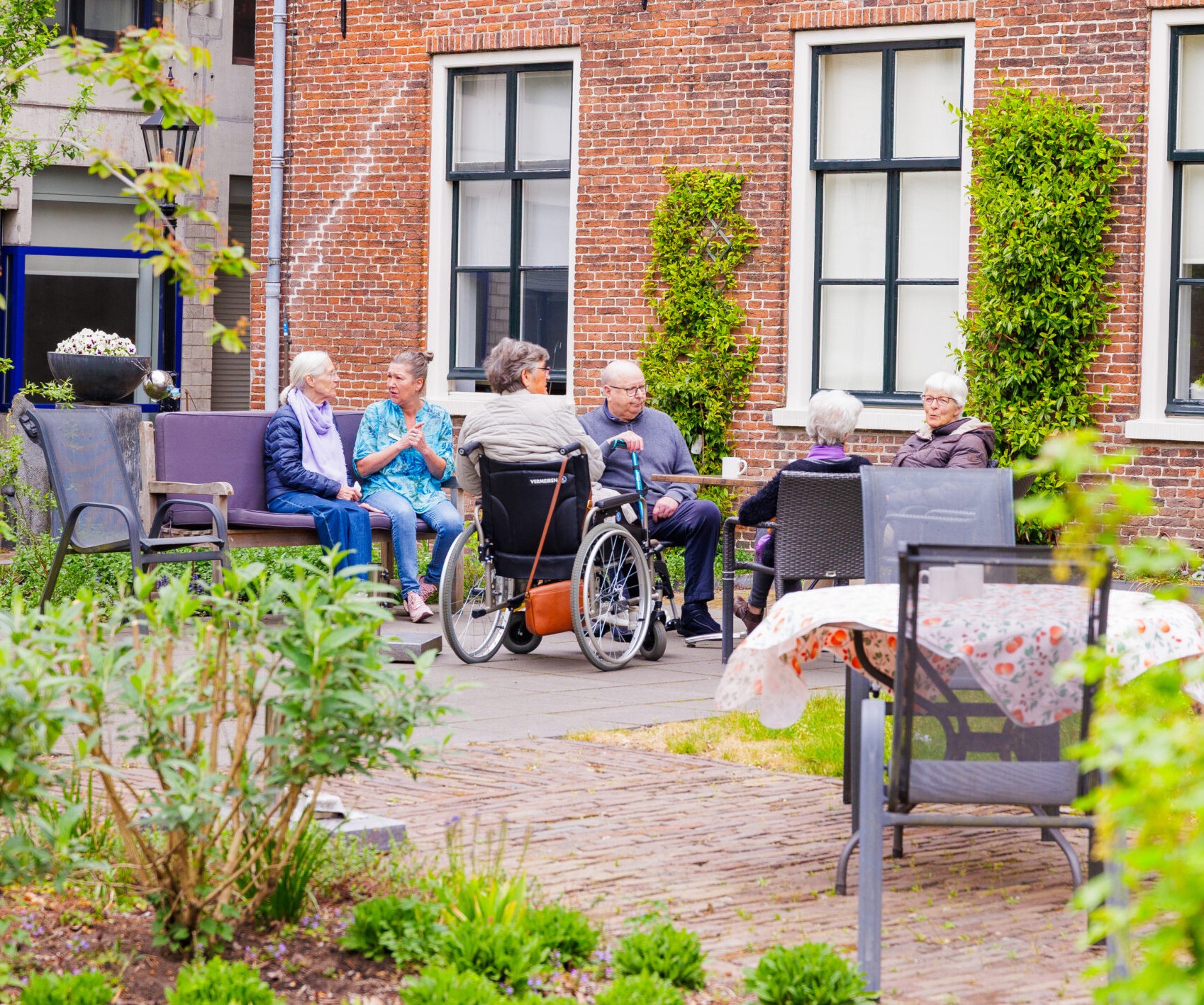Een groep ouderen en een zorgmedewerker zitten gezellig samen in een groene, beschutte binnentuin bij een historisch bakstenen gebouw.