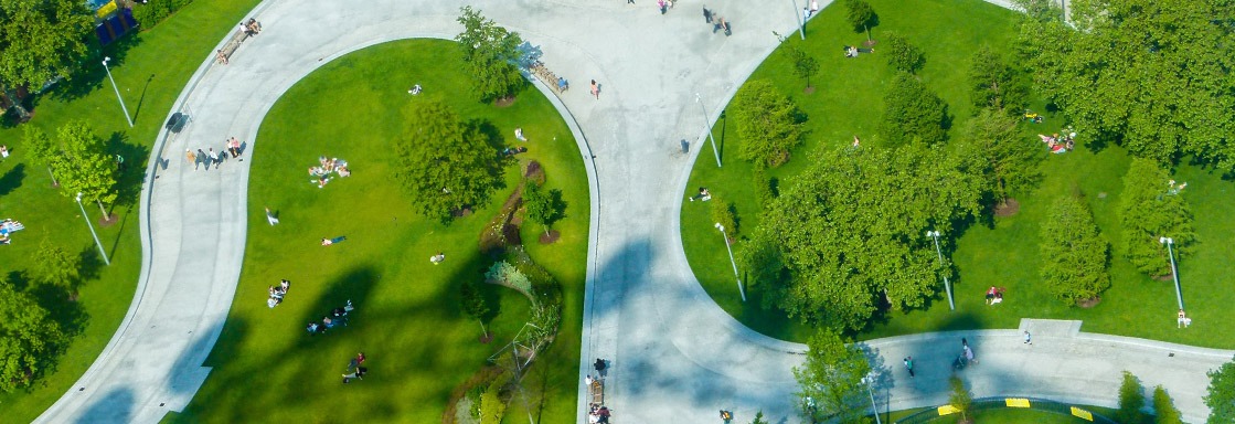 Een vogelvluchtperspectief van een modern, groen stadspark met kronkelende, brede betonnen wandelpaden, grasvelden waar mensen op ontspannen, bomen en lantaarnpalen.